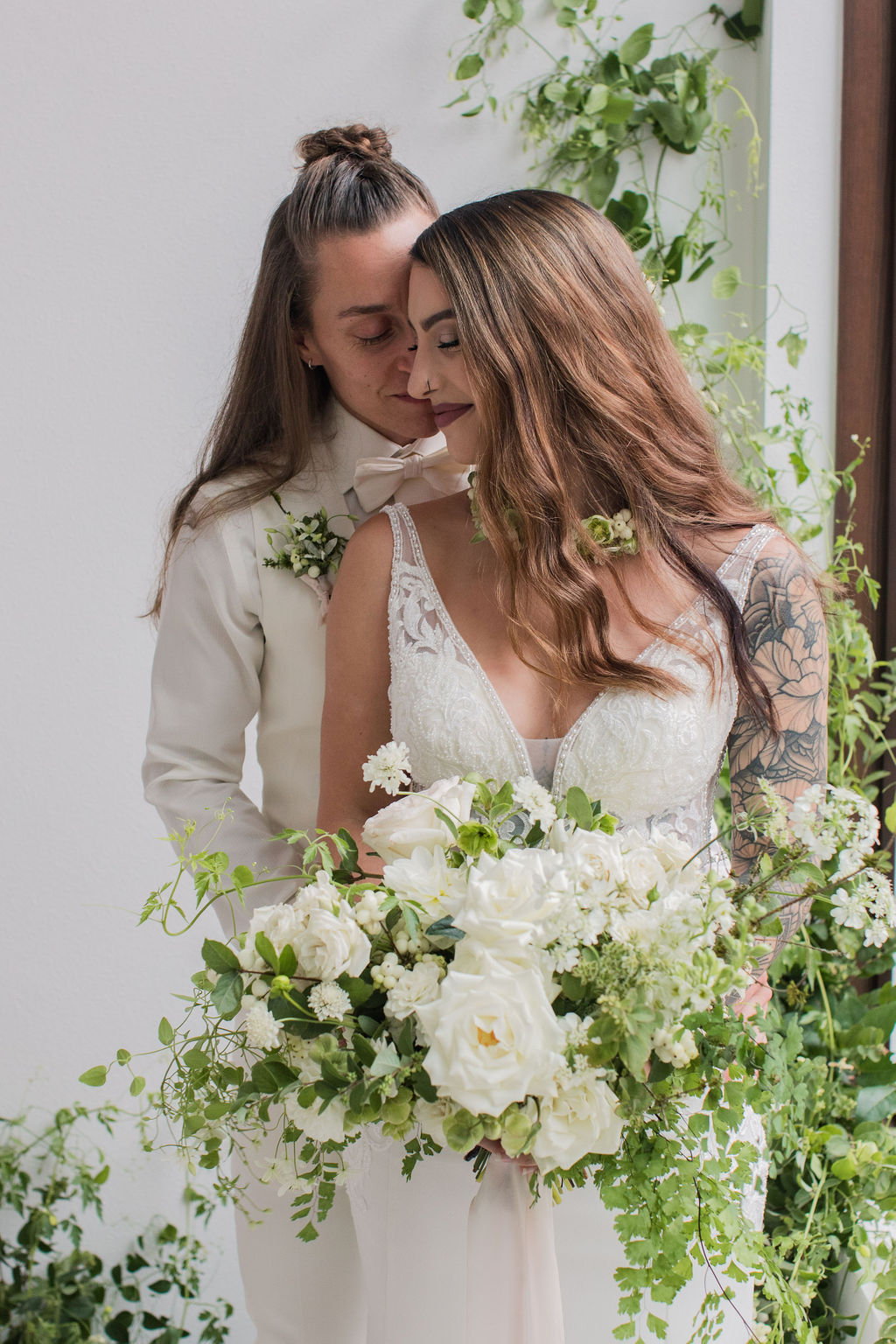 two brides holding a bouquet in front of a greenery installation, smiling