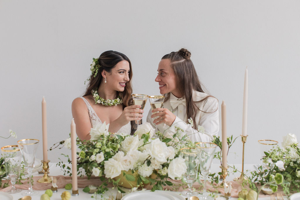 two brides clinking champagne glasses at a wedding sweetheart table covered in white and green floral arrangements, a neutral table runner, neutral candles, and white and gold table settings