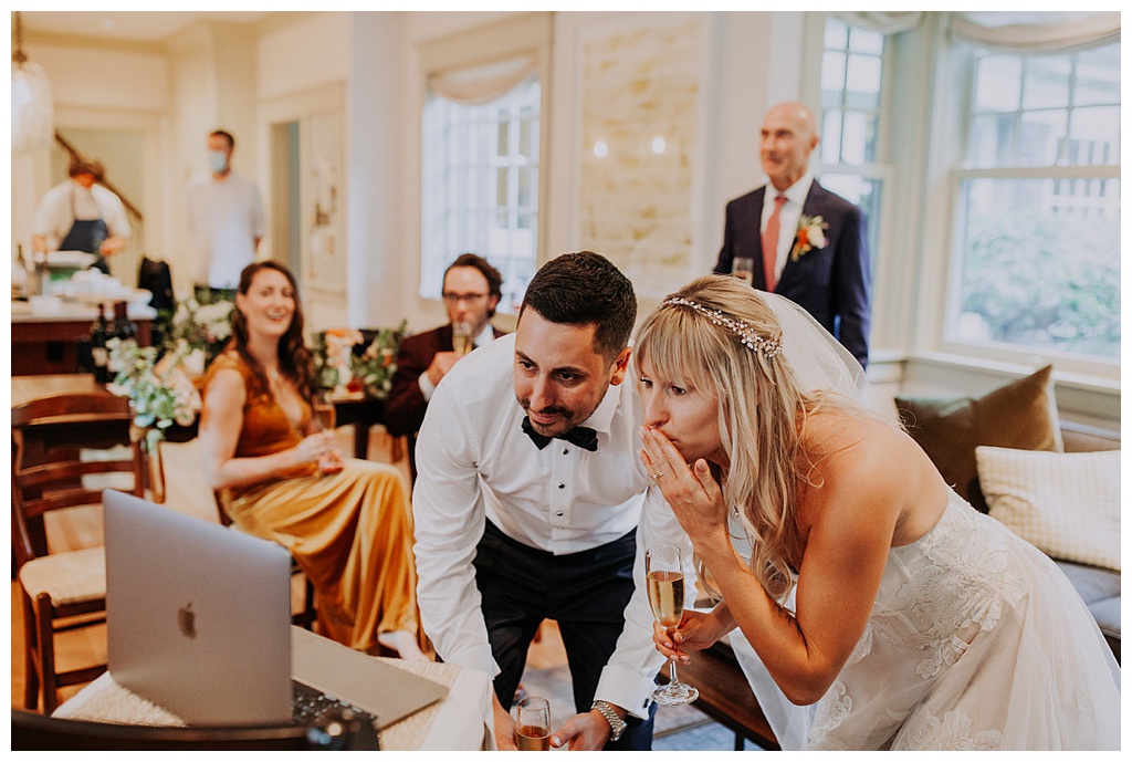 a bride and groom looking at a laptop and blowing kisses with their family in the background